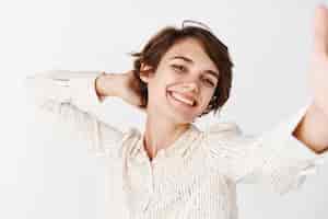 Бесплатное фото Откровенная счастливая женщина позирует для селфи со смартфоном и фотографирует себя с веселой улыбкой, беззаботно стоящей на белом фоне
