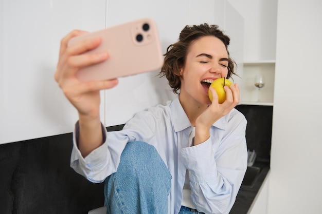 Foto gratuita una giovane donna candida e felice che morde una mela per un selfie scatta una foto sullo smartphone mentre mangia frutta