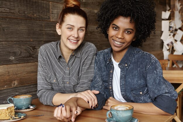 Candid indoor shot of cute homosexual couple holding hands and looking at camera with happy smiles