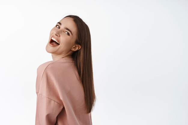 Candid people. Happy young emotional girl, turn face at camera and posing for shot, smiling and laughing carefree, standing against white background