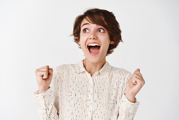 Free photo candid portrait of young happy woman winning and triumphing looking left at empty space and scream yes with joy achieve goal or prize celebrating victory standing on white background