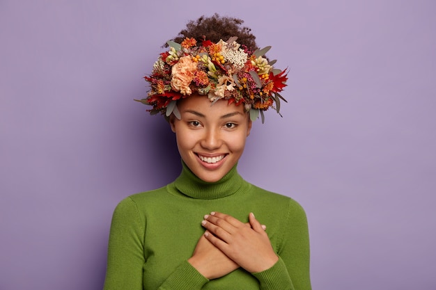 Candid shot of attractive smiling woman feels thankful, ouches chest gratefully, wears autumn wreath, poses indoor in casual wear