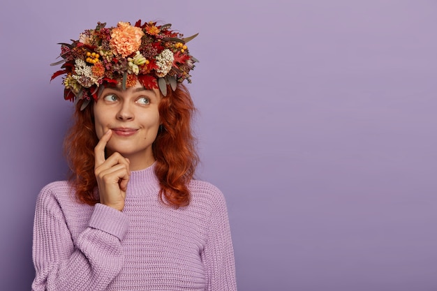 Candid shot of beautiful redhead woman has dreamy expression, keeps finger near lips, being deep in thoughts, wears plant wreath