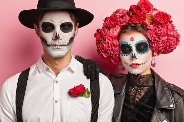 Free photo candid shot of calm woman and man zombie with eyes closed, have artistic makeup, traditional holiday costumes, celebrate day of dead, have scary look, isolated over pink background.