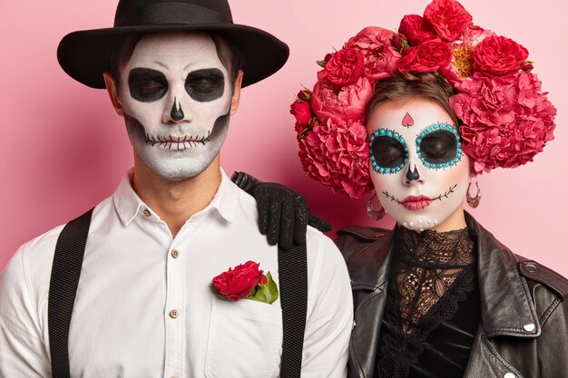 Candid shot of calm woman and man zombie with eyes closed, have artistic makeup, traditional holiday costumes, celebrate day of dead, have scary look, isolated over pink background.