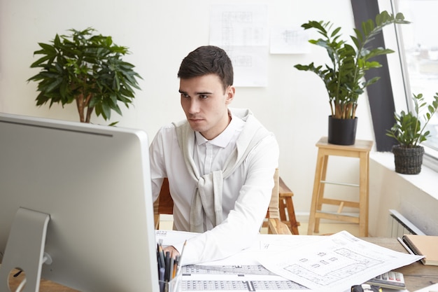 Candid shot of concentrated young brunette man engineer dressed formally working on generic computer, having serious look, sitting in his modern office, surrounded with papers and blueprints