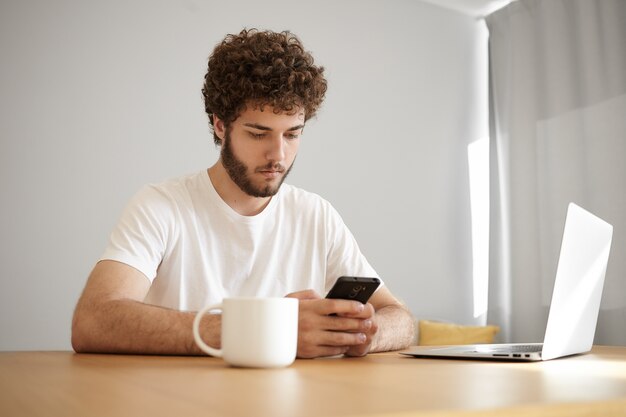 그의 직장에 앉아 멀리 노트북을 사용하고 커피를 마시는 동안 휴대 전화에 문자 메시지를 입력하는 흰색 티셔츠에 집중된 젊은 수염 난 남성 프리랜서의 솔직한 샷