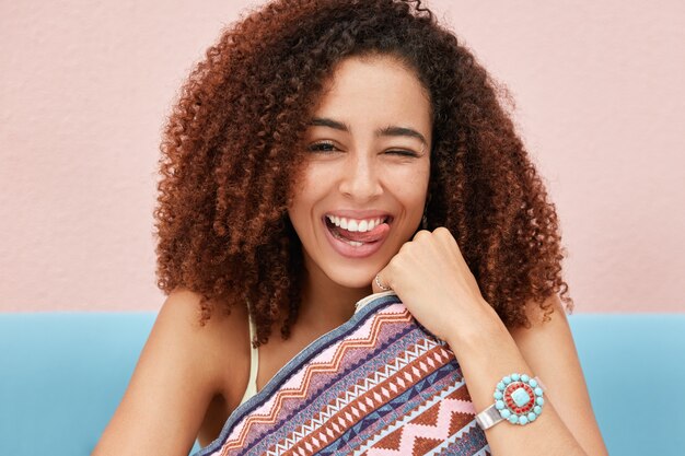 Candid shot of funny dark skinned pretty woman foolishes, shows tongue and blinks eye, holds cushion, has fun together with friends during party meet for entertaining.