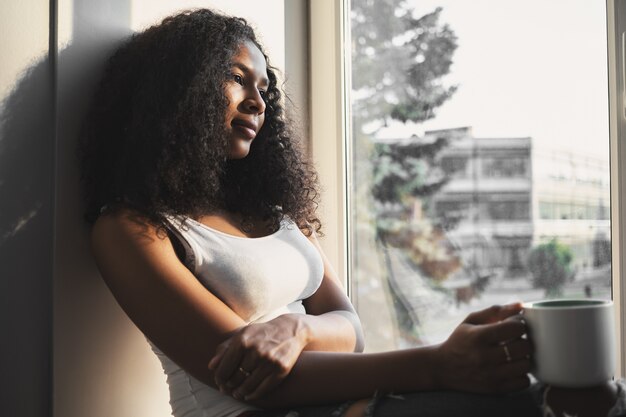 Candid shot of gorgeous charming young dark skinned woman with voluminous hairdo relaxing indoors alone, sitting on windowsill with cup of coffee, having thoughtful dreamy facial expression