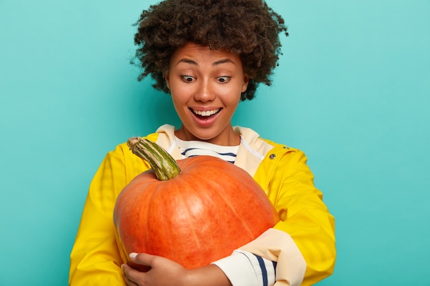 Candid shot of happy mixed race young woman embraces big pumpkin, dressed in yellow raincoat, pleased to grow huge vegetable in own garden, models against blue background. Autumn decoration.