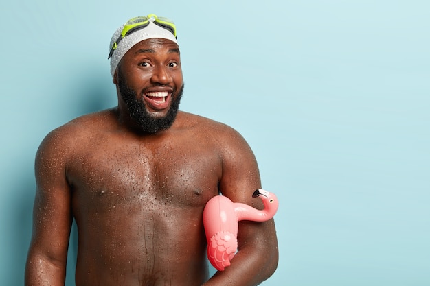 Free photo candid shot of joyous dark skinned guy receives splash of water on naked body, wears goggles, carries swiming ring