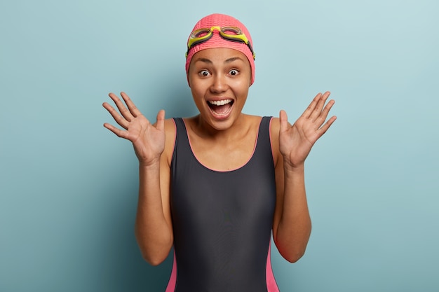 Candid shot of overemotive African American woman raises palms, has happy reaction, wears black swimsuit