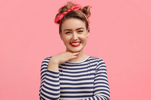 Candid shot of pleasant looking female with make up, has positive expression, keeps hand under chin, wears striped sweater and stylish headband