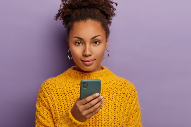 Candid shot of serious millennial woman with curly dark hair, uses mobile phone, looks directly at camera, wears yellow clothes
