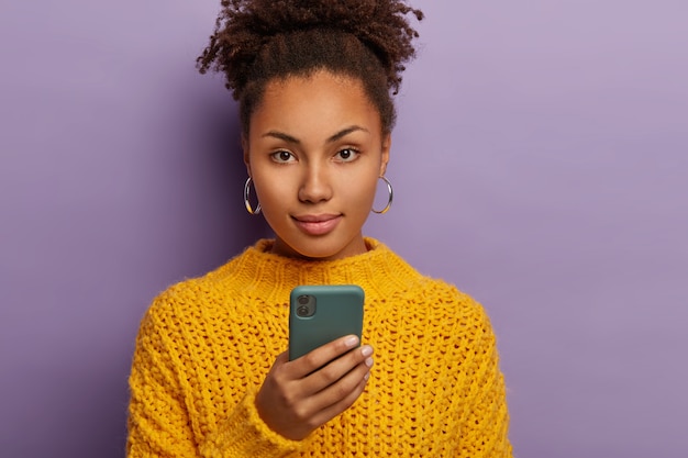 Candid shot of serious millennial woman with curly dark hair, uses mobile phone, looks directly at camera, wears yellow clothes