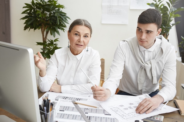 Foto gratuita candido colpo di elegante esperto architetto femmina senior dai capelli grigi che punta allo schermo del computer, spiegando il punto di vista al suo giovane collega maschio mentre lavorava insieme su un nuovo progetto abitativo,