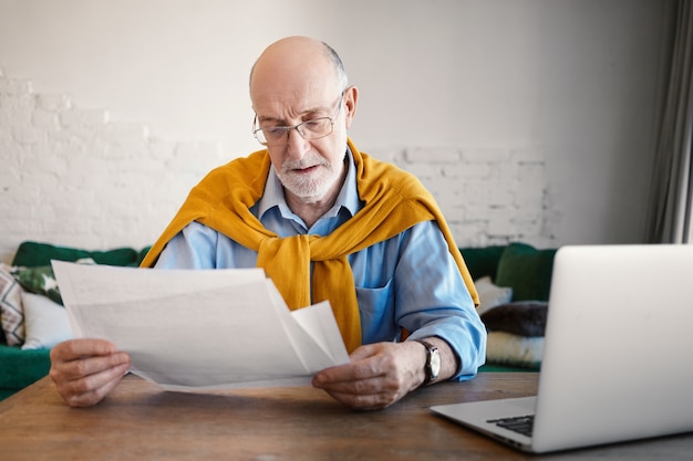 Candid shot of stylish experienced senior bearded engineer holding technical drawings, checking measurements and calculations of engineering project, having focused concentrated facial expression