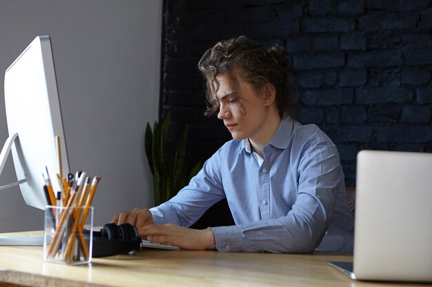 現代のオフィスでパソコンとラップトップを使用して、巻き毛を集めたスタイリッシュな若い男性プログラマーの率直なショット。仕事、ビジネス、キャリア、職業