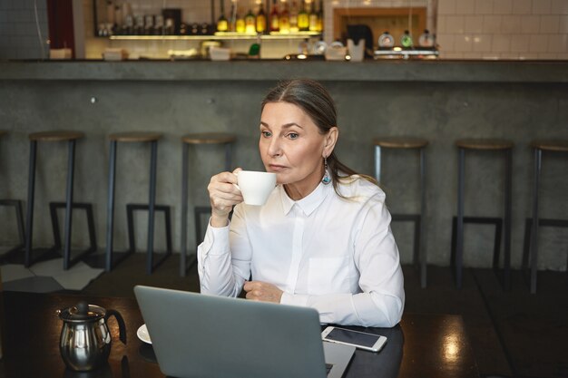 Откровенный снимок вдумчивой зрелой бизнес-леди в формальной рубашке, наслаждающейся кофе во время обеда, сидящей в кафе с универсальным ноутбуком и сотовым телефоном с пустым экраном на столе. Бизнес, возраст и технологии