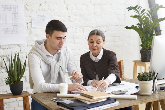 Candid shot of two coworkers brainstorming in office: middle aged female chief engineer proving her point of view to her young brunette male colleague, pointing at blueprint in front of her on desk