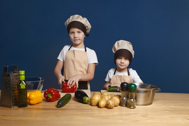 Scatto schietto di due bambini maschi che indossano cappelli da cuoco e grembiuli che fanno il pranzo insieme al tavolo della cucina