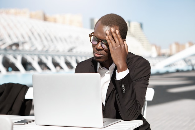 Откровенный снимок несчастного молодого афроамериканского менеджера, который чувствует себя уставшим и расстроенным, сидит в городском кафе с обычным ноутбуком, трогает голову, пытается сосредоточиться на работе, выглядит измотанным