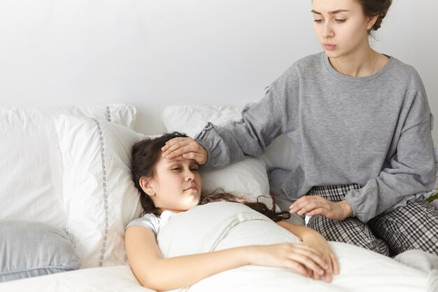 Candid shot of worried upset young woman sitting in bedroom, holdings hand on her sick daughter's forehead,