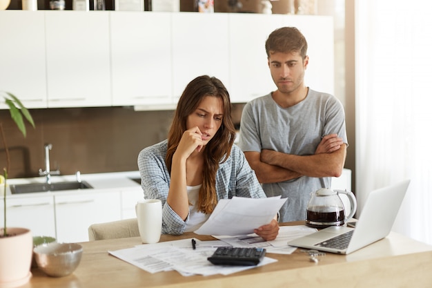 Candid shot of young American man and woman dressed casually feeling stressed while managing finances in kitchen together, calculating expenses, paying utility bills online on laptop computer