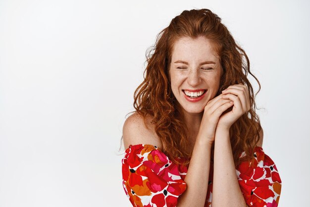 Candid smiling girl with curly red hair laughing and grinning with closed eyes holding hands near face standing in dress against white background