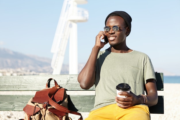 Candid urban shot of cheerful dark-skinned student sittingoutdoors on wooden bench with backpack and having nice phone conversation with friend on mobile, smiling happily, enjoying takeaway coffee