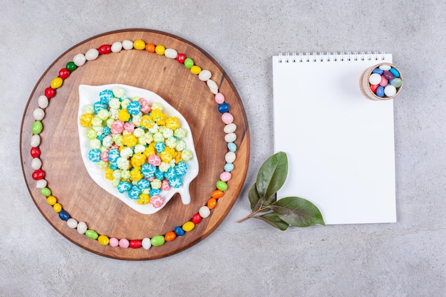 Candies in and around an ornate platter on a wooden tray next to a notepad adorned with leaves on marble background. High quality photo