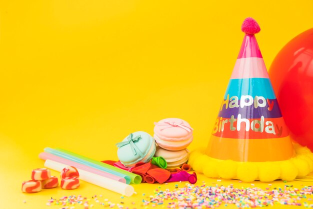 Candies; straw; deflated balloon; macarons and paper hat on yellow background