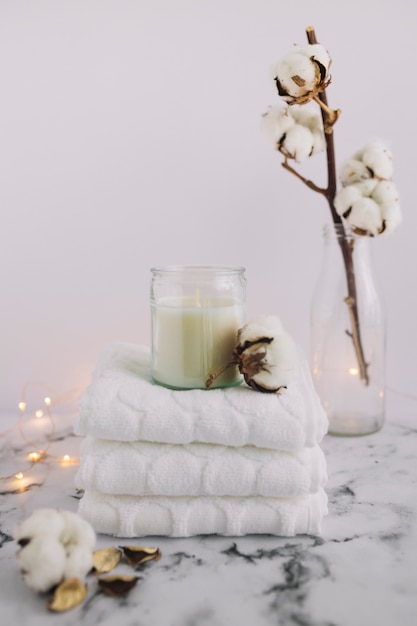 Candle in candleholder with stacked napkins near cotton twig and lighting equipments on marble surface