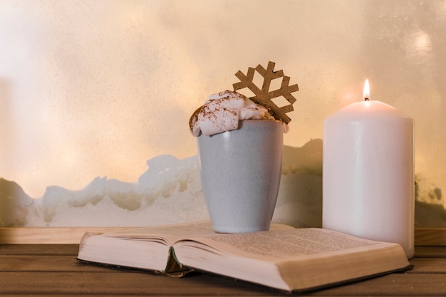 Candle near book and cup with toy snowflake on wood board near heap of snow through window