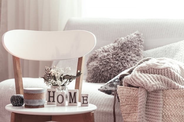 Candles, a vase with flowers with wooden letters of the home on wooden white chair. Sofa and wicker basket with cushions in the background.