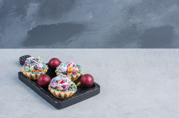 Candy topped cupcakes and baubles in a black tray on marble surface