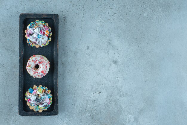 Candy-topped cupcakes and donuts on a black tray on marble surface