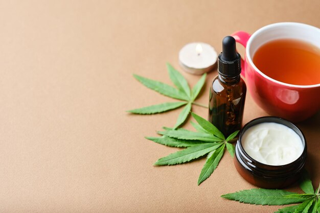 Cannabis cosmetic oil cream in jar bottle herbal and a green plant leaf Natural cosmetic on brown background Flat lay copy space