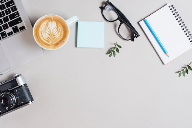 Cappuccino coffee cup; laptop; retro camera; adhesive notepad; eyeglasses and pencil on spiral notepad against white backdrop