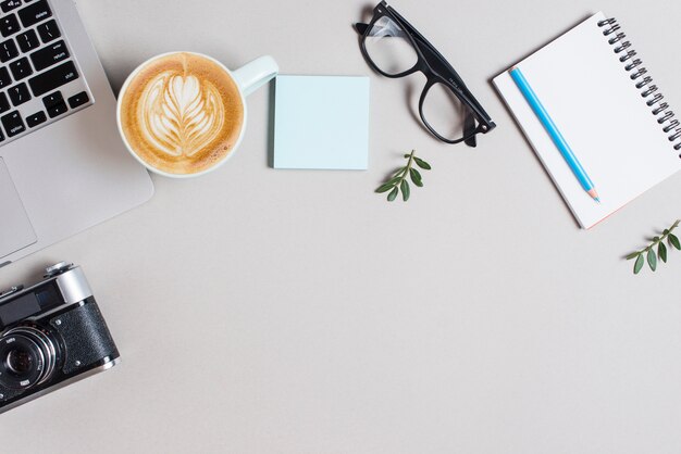 Cappuccino coffee cup; laptop; retro camera; adhesive notepad; eyeglasses and pencil on spiral notepad against white backdrop