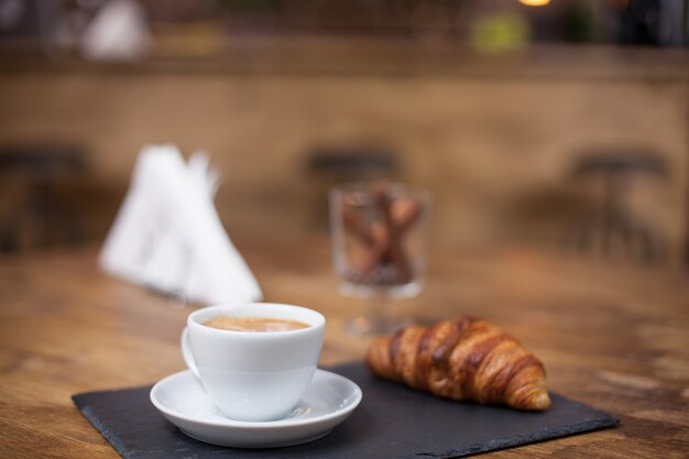 Cappuccino coffee in a white cup on a wooden table next to a delicious croissant. Tasty snak. Vintage coffee shop.