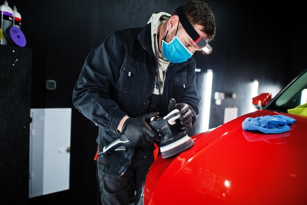 Car detailing concept Man in face mask with orbital polisher in repair shop polishing orange suv car