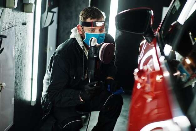 Car detailing concept Man in face mask with orbital polisher in repair shop polishing orange suv car