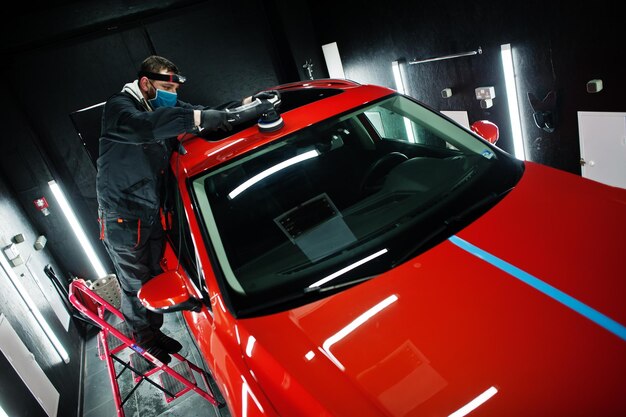 Концепция детализации автомобиля Человек в маске с орбитальной полировальной машиной в ремонтной мастерской полирует крышу оранжевого внедорожника