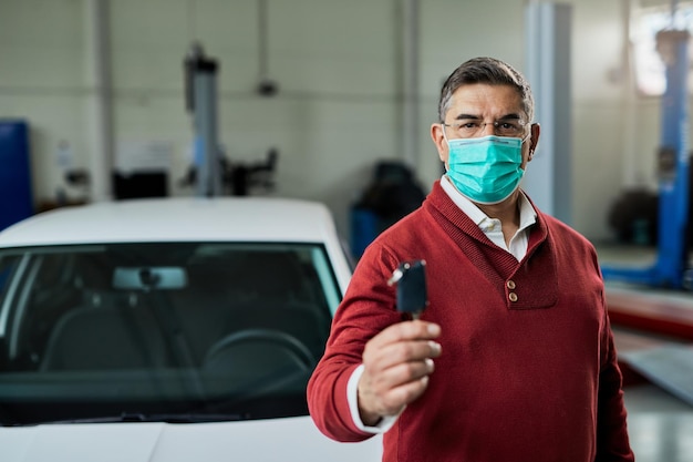コロナウイルスの流行のために保護フェイスマスクを着用しながらワークショップで彼の修理された車のキーを保持している車の所有者