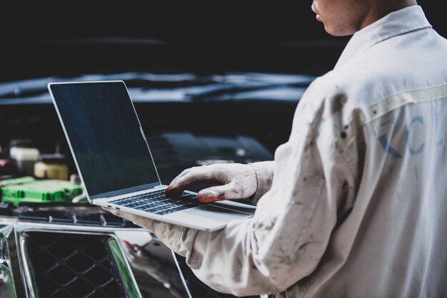 Car repairman wearing a white uniform standing and holding a wrench that is an essential tool for a mechanic with laptop checking engine