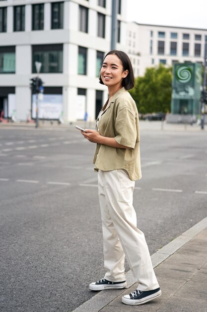 Car sharing and technology young asian woman waiting for taxi near road holding smartphone order car