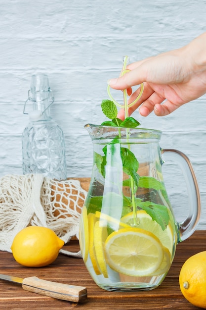 Carafe of lemon juice with wooden knife, lemons, hand holding leaves side view on a wooden and white surface