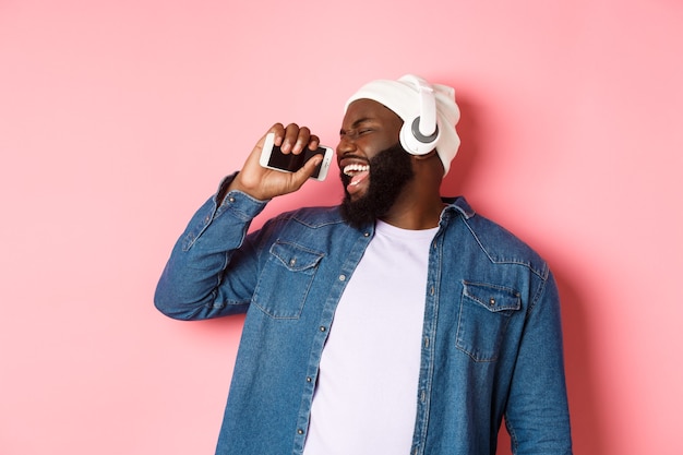 のんきなアフリカ系アメリカ人の男性がヘッドフォンで音楽を聴き、マイクとして携帯電話で歌い、ピンクの背景の上に立っています