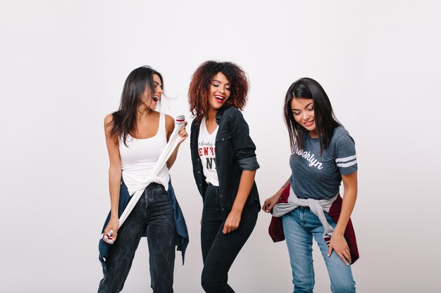 Carefree african girl having fun with fashionable friends smiling. Excited black female model standing with eyes closed while brunette ladies dancing beside.